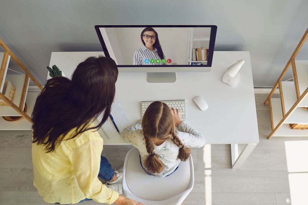Woman and child at desk, video call with teacher on screen.