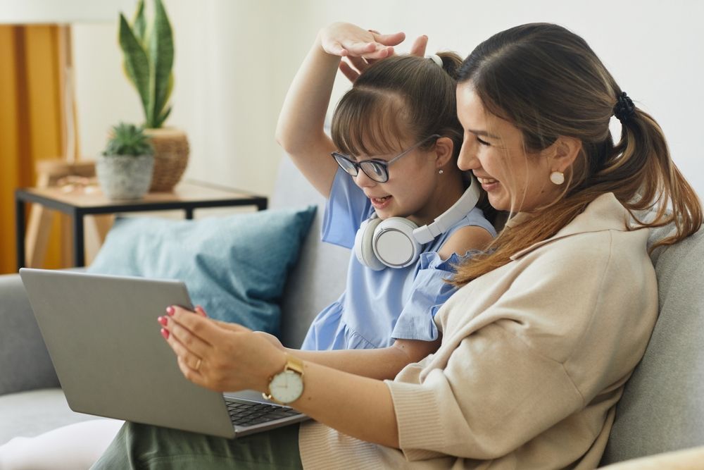 Woman and child with headphones on looking at a laptop together, smiling. Living room setting.