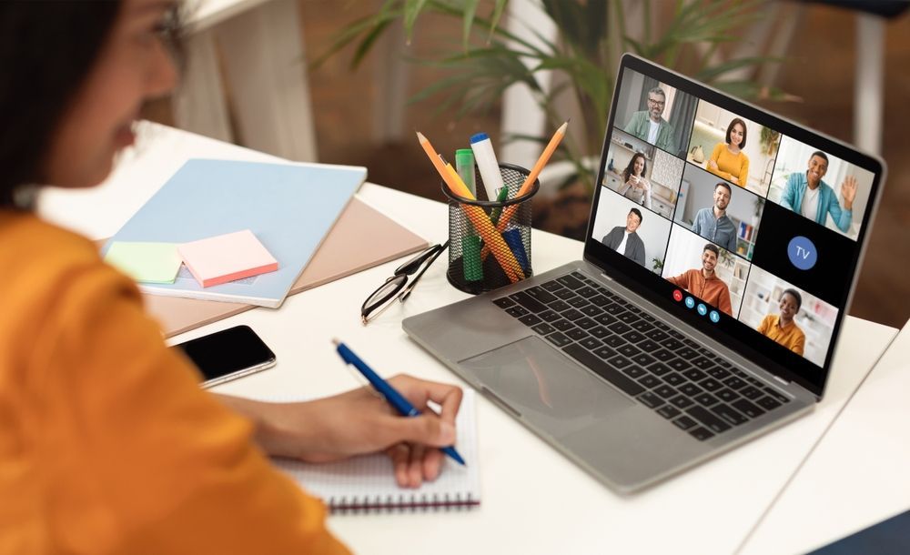 Woman taking notes during a video conference on a laptop, with colleagues visible in individual screen tiles.
