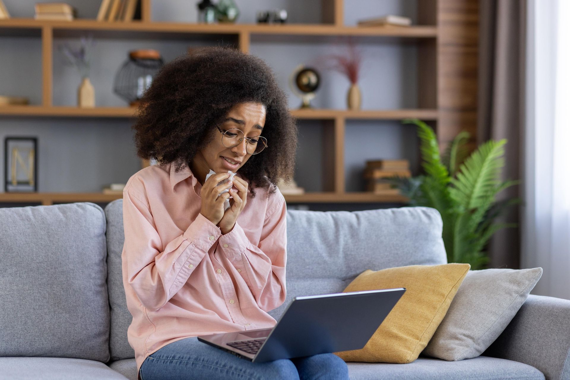Woman on couch, looking at laptop with sad expression, holding tissue; living room.