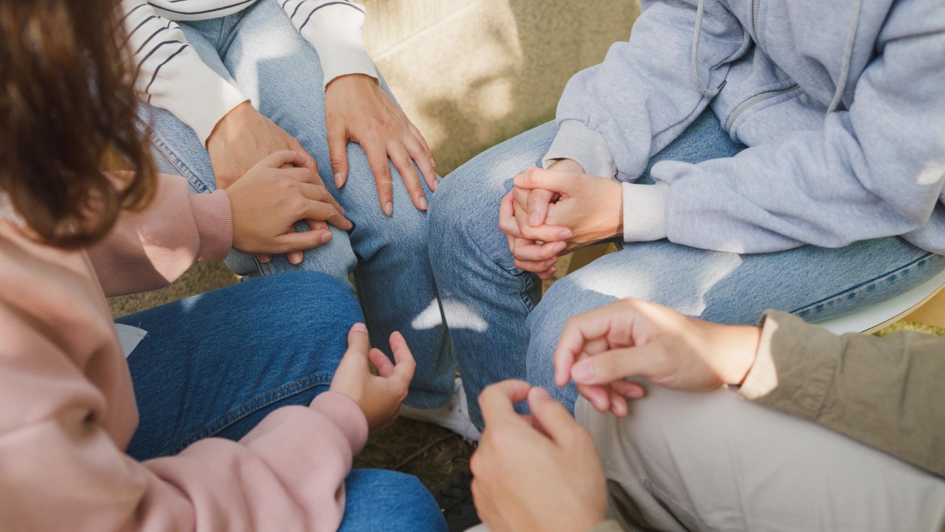 Hands of several people clasped together in a circle, on laps, in a sunlit room.