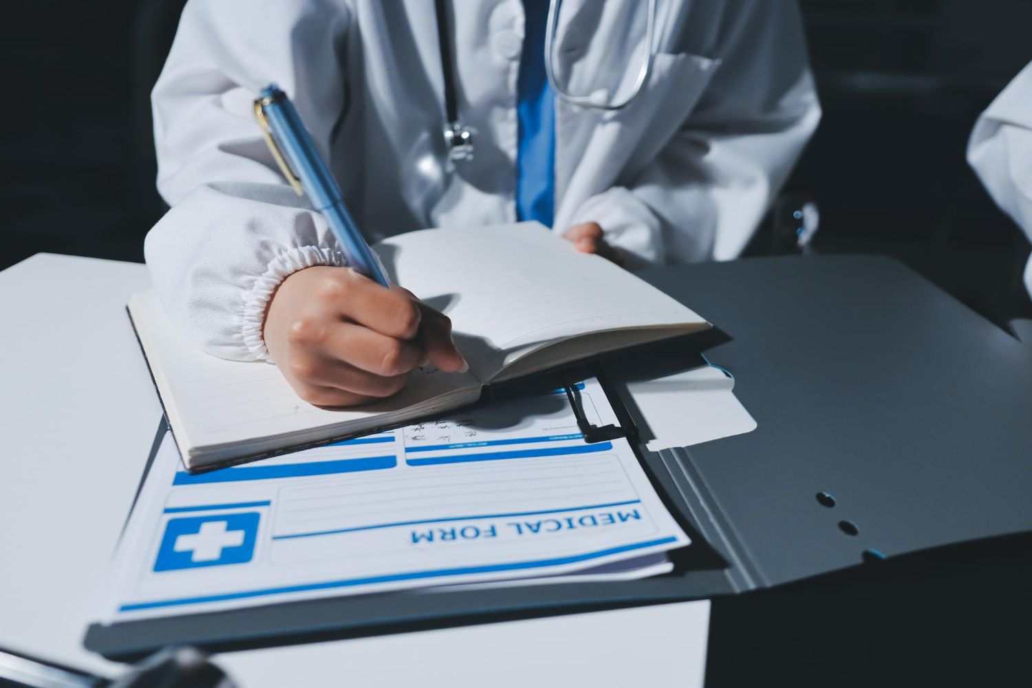 Person writing in a notebook at a table with a laptop and another person using a phone.