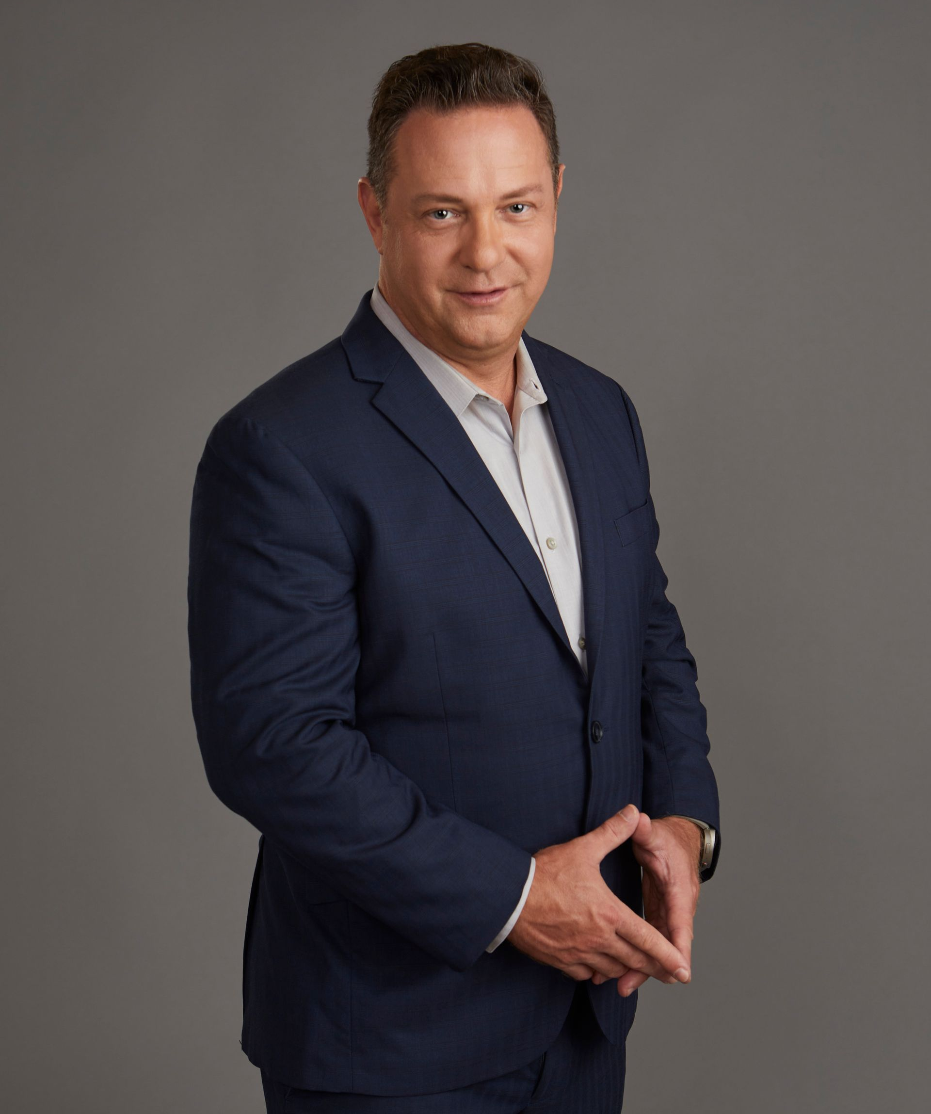Man with shaved head and goatee smiles broadly, wearing a black shirt, set against green foliage.