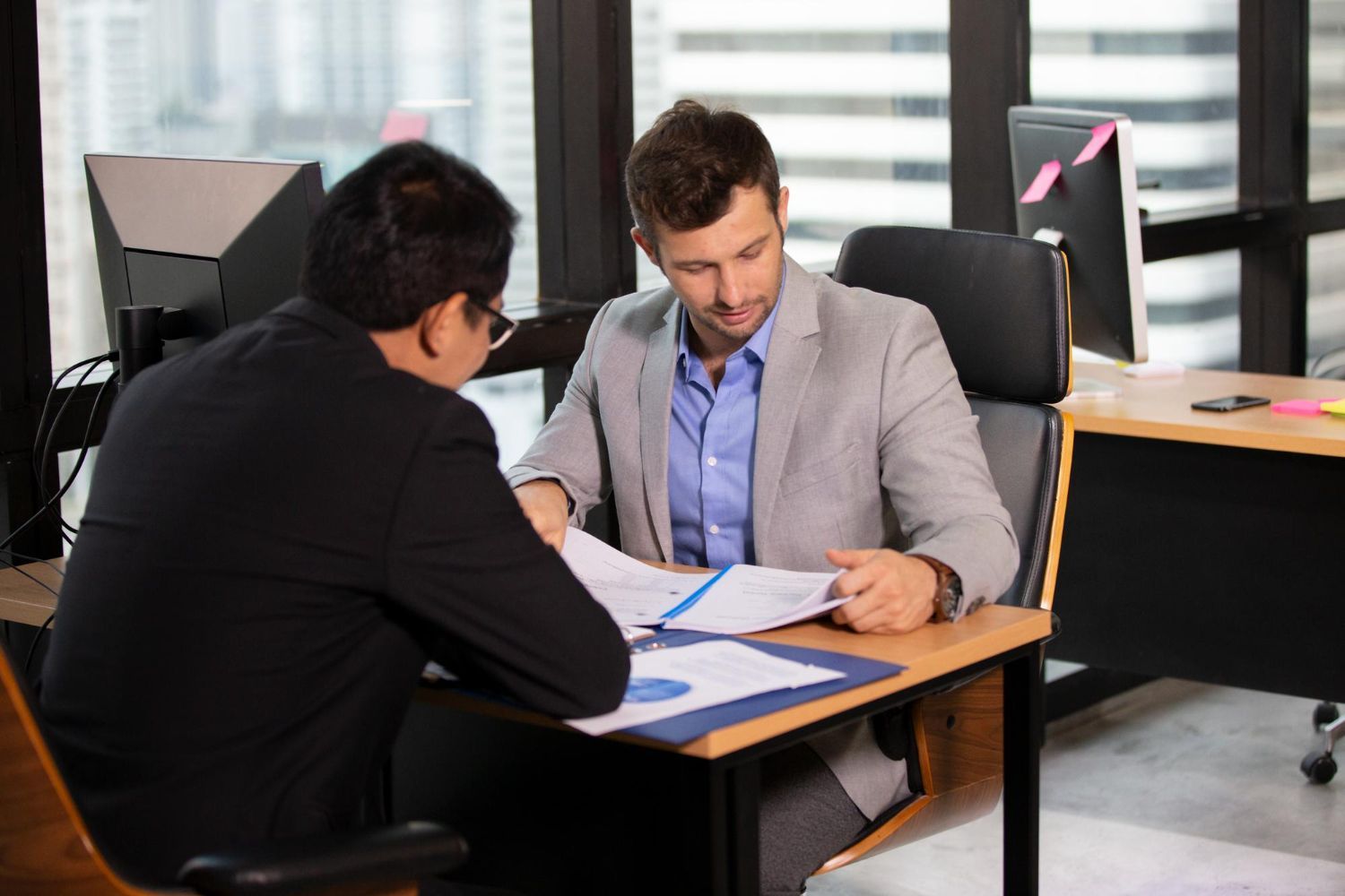 Person writing in a notebook at a table with a laptop and another person using a phone.