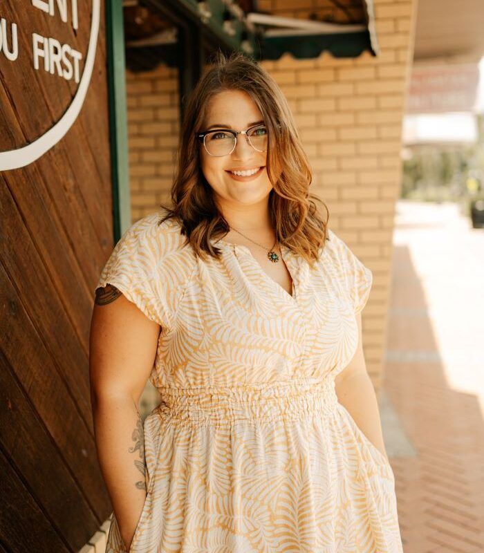 Woman with glasses in a yellow dress, smiling, standing in front of a building entrance.