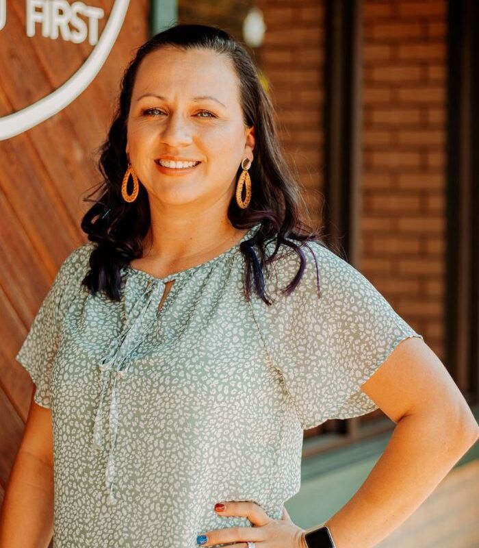 Woman with dark hair, smiles, wearing green patterned blouse, gold earrings, hand on hip.