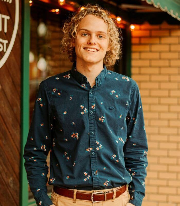 Man with curly blonde hair smiles, wearing a blue floral shirt and tan pants outside a shop.