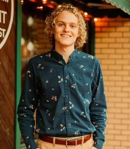 Young man with curly blonde hair smiles, wearing a blue floral button-up shirt, tan pants, and a brown belt.