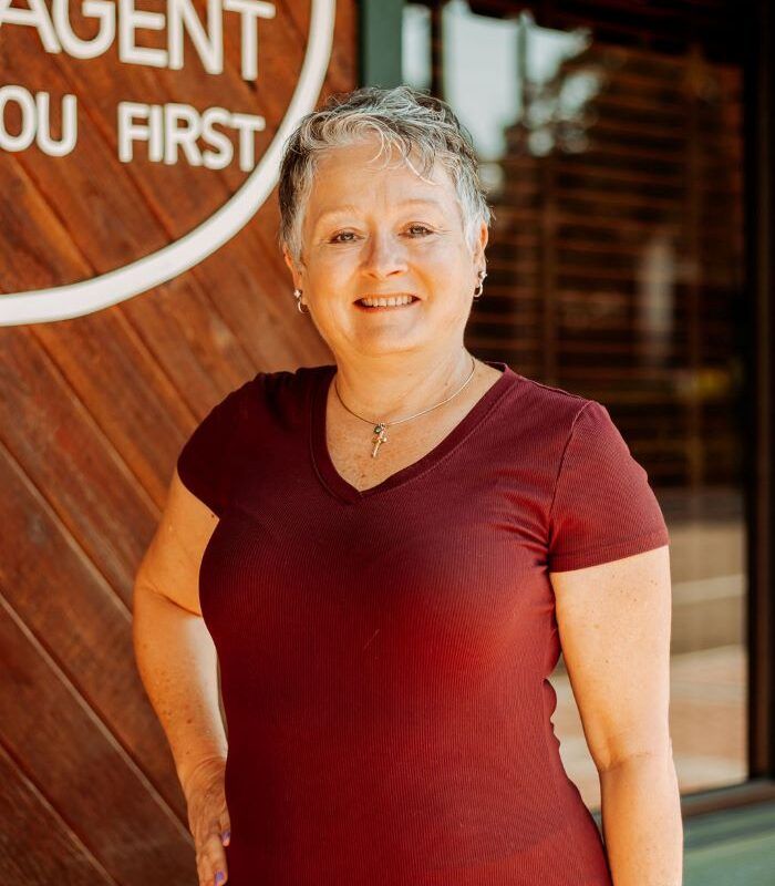 Woman with short gray hair, smiling, wearing a burgundy top, standing near a wooden door.