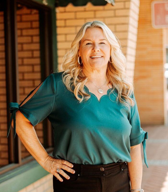 Woman with blonde hair and green top, standing with hand on hip in front of brick building.