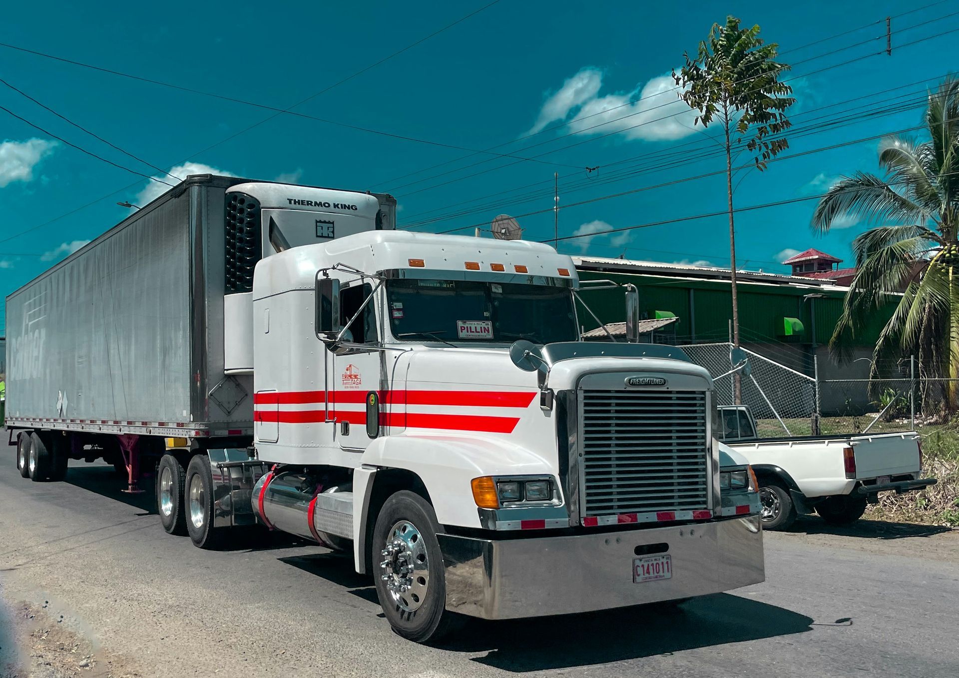 White semi-truck with red stripes on a road, carrying a refrigerated trailer, parked in front of a building.