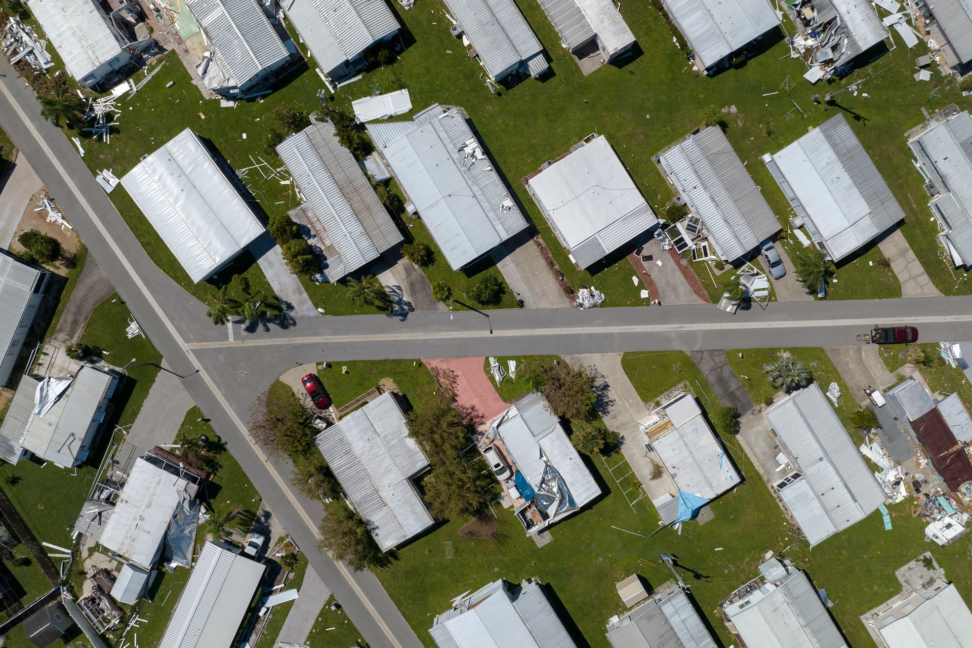 Aerial view of damaged mobile homes after a storm, with torn roofs and debris scattered across the green lawns.