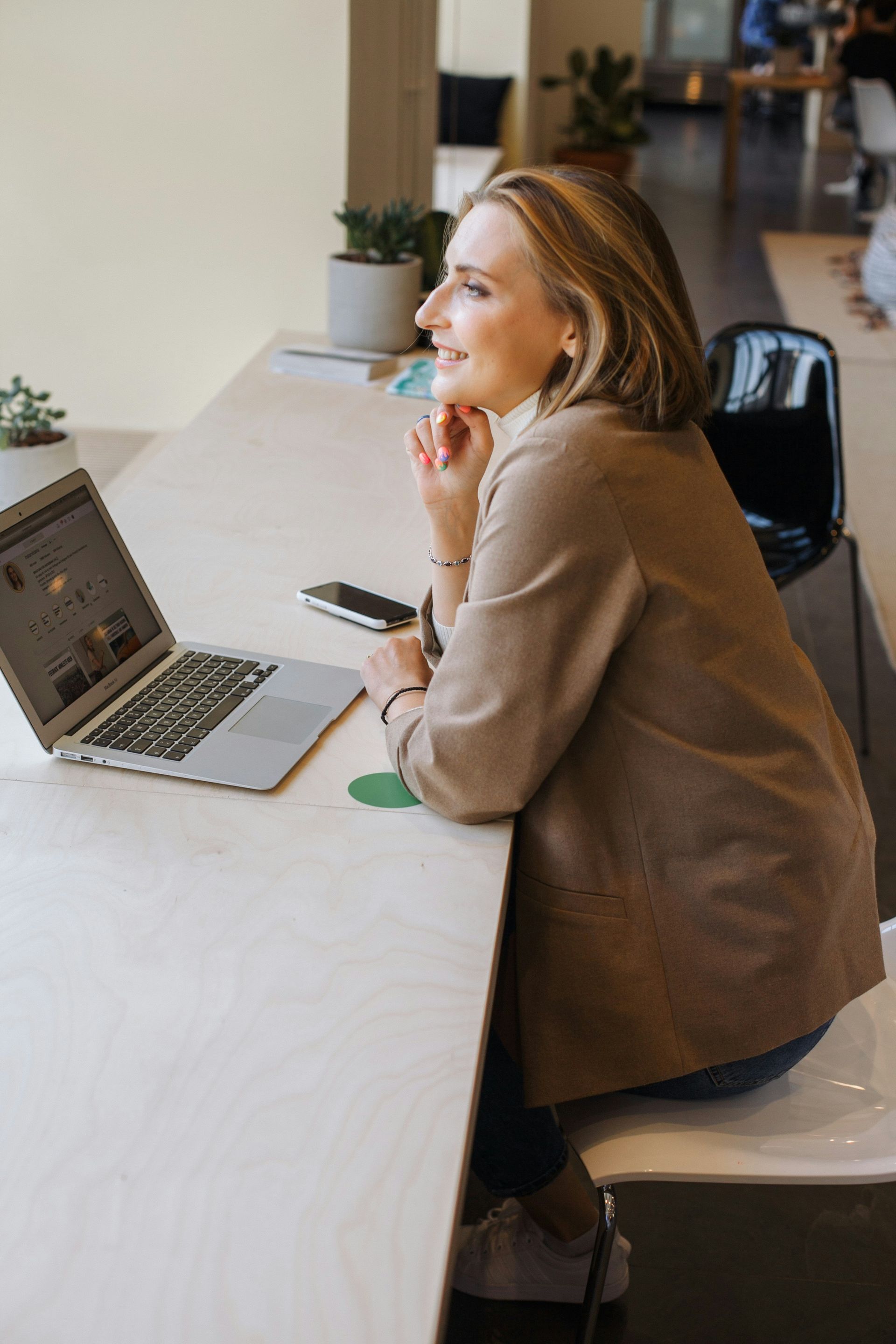 Woman seated, smiling at a window while working on a laptop in a light-filled office space.