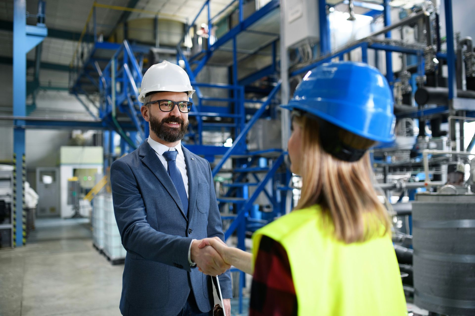 Man in suit shaking hands with woman in hard hat, factory setting.