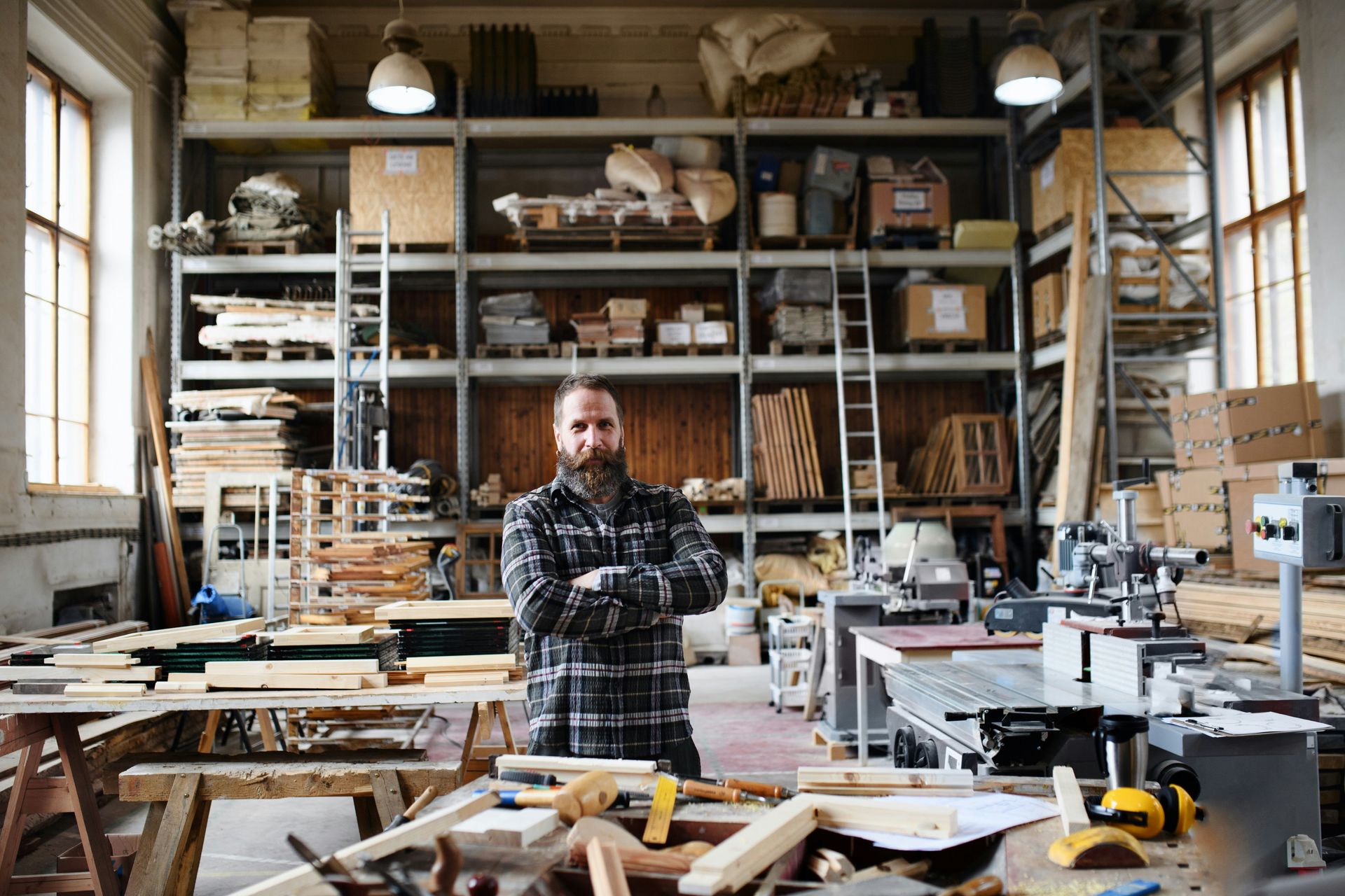 Man in plaid shirt stands in a cluttered wood workshop, arms crossed, surrounded by tools and wood.