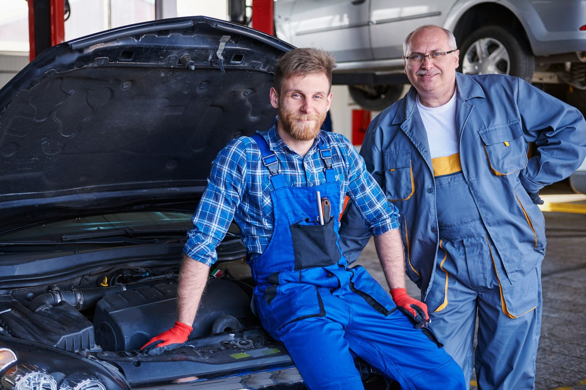 Two mechanics in a garage, one leaning on a car with the hood up; both wearing work overalls.