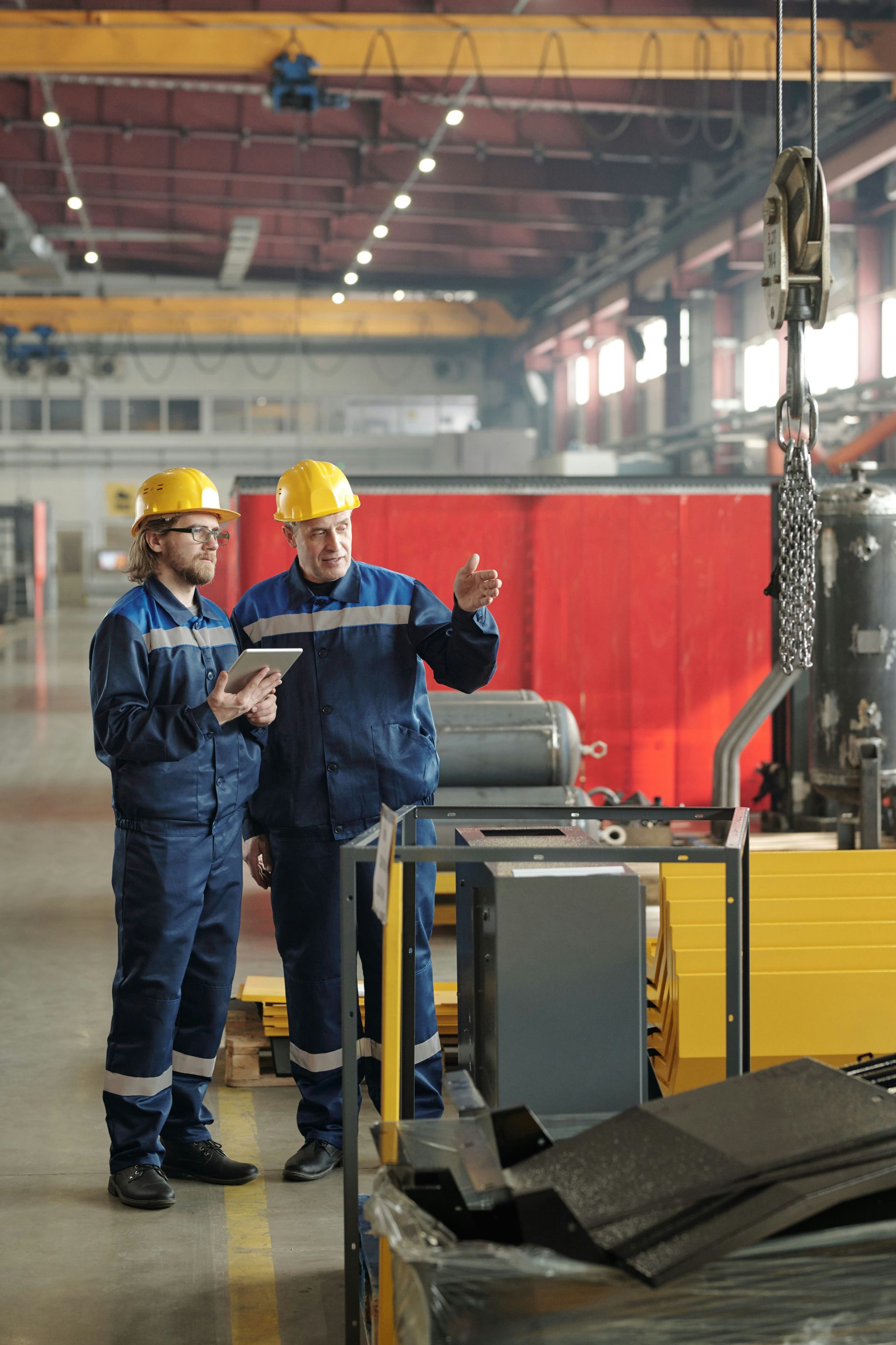 Two factory workers in blue coveralls and yellow hardhats reviewing a tablet in a factory setting.