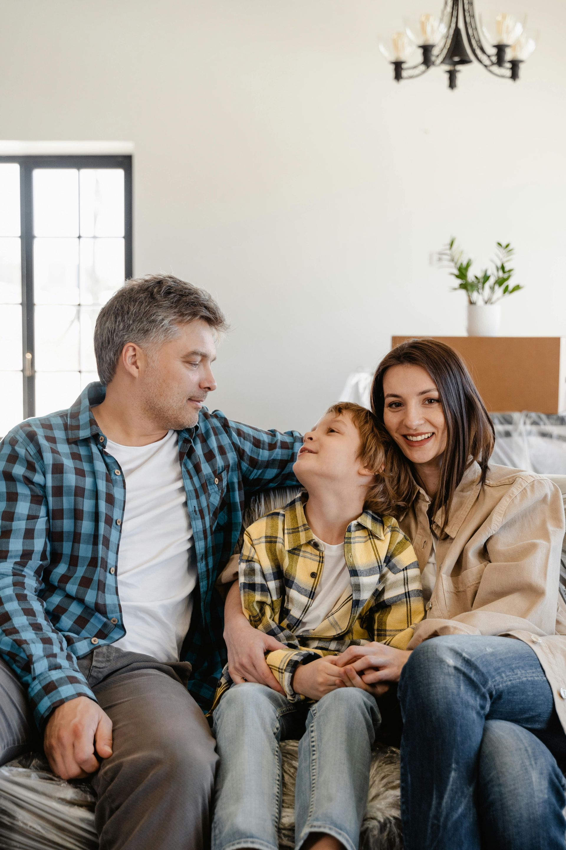 Family of three sitting on couch, smiling. Father in plaid shirt, son in yellow, mother in jeans.