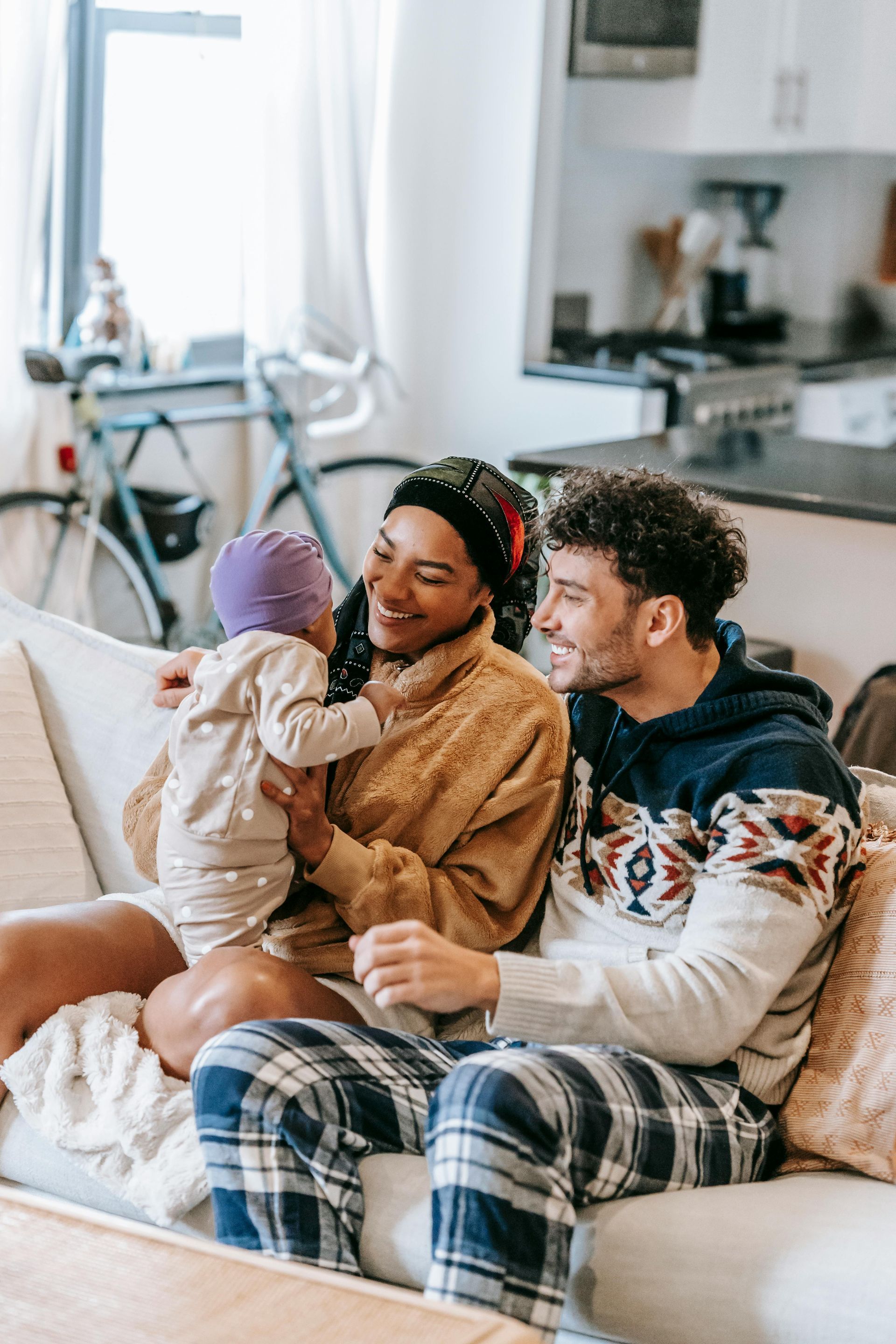 Family of three on a couch, woman holding child, man smiles. Cozy setting with a bike in the background.