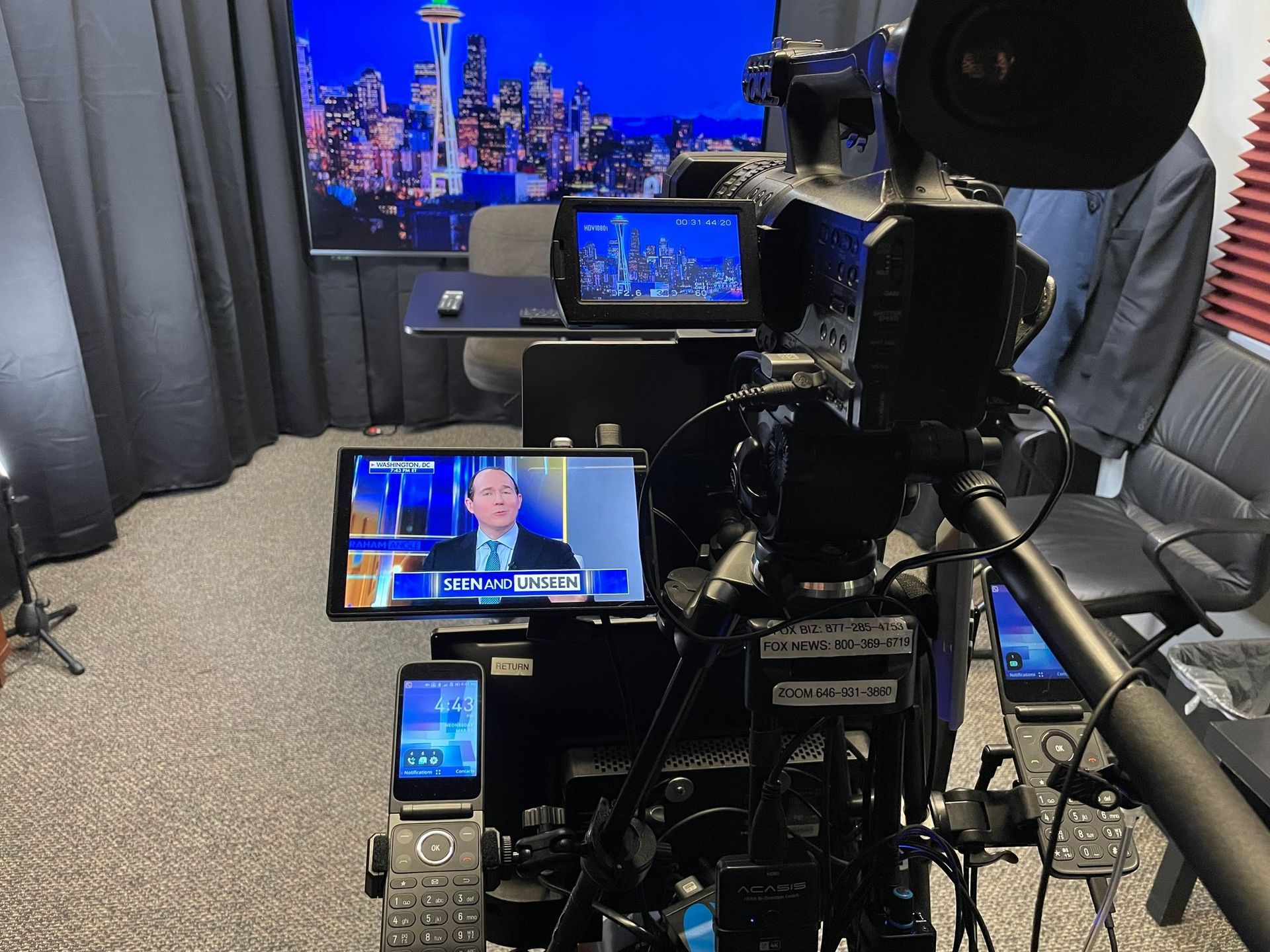 Video camera pointed at a home studio desk with multiple monitors showing a news broadcast and city skyline background.