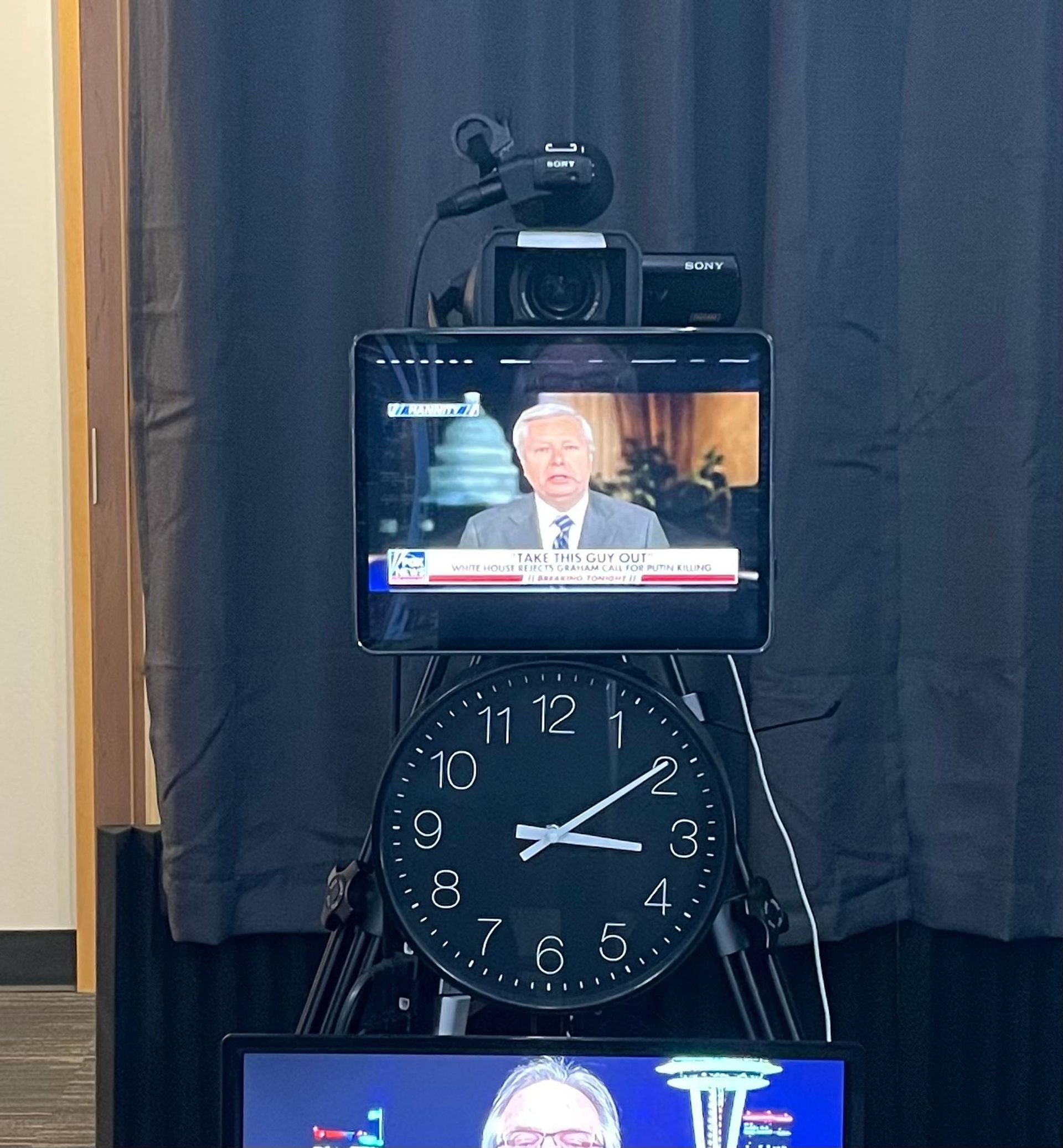 TV screen atop a large black clock in front of dark curtains, with a smaller screen visible below