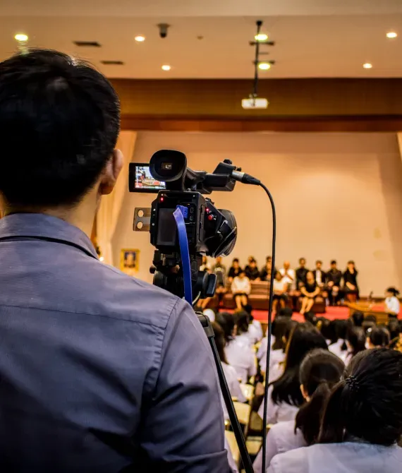 Videographer filming a panel discussion in a conference hall, with audience seated in the foreground.