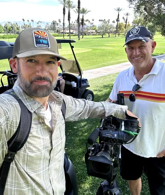 Two men smiling beside a golf cart on a sunny golf course, one holding a camera rig.