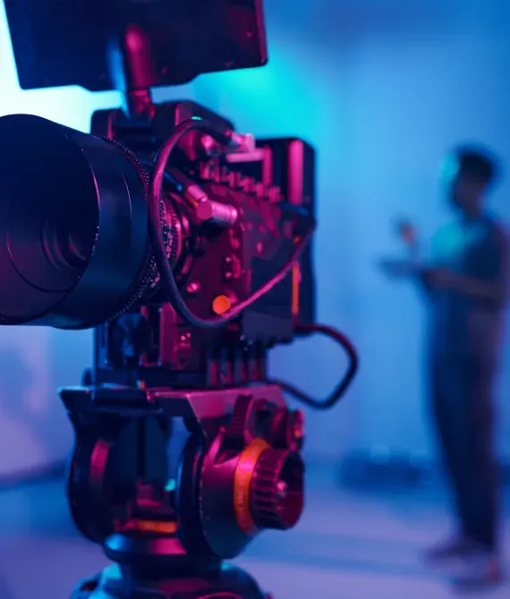 Close-up of a professional camera with pink and blue lighting, filming a blurred person in the background