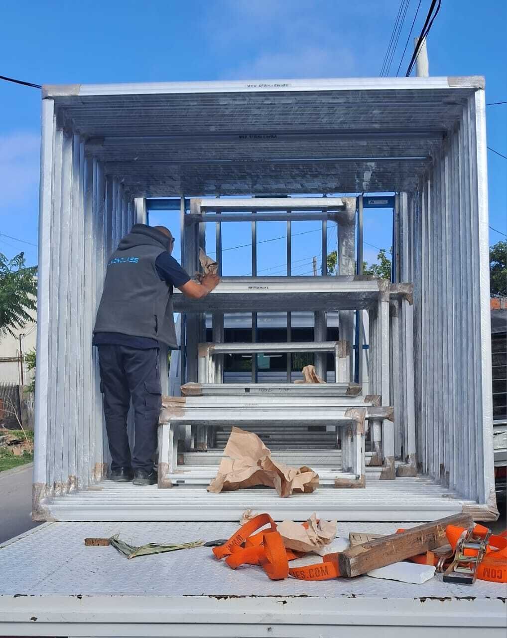 Persona cargando ventanas en un camión. Cielo azul, plataforma metálica. Trabajador dentro, con chaleco.