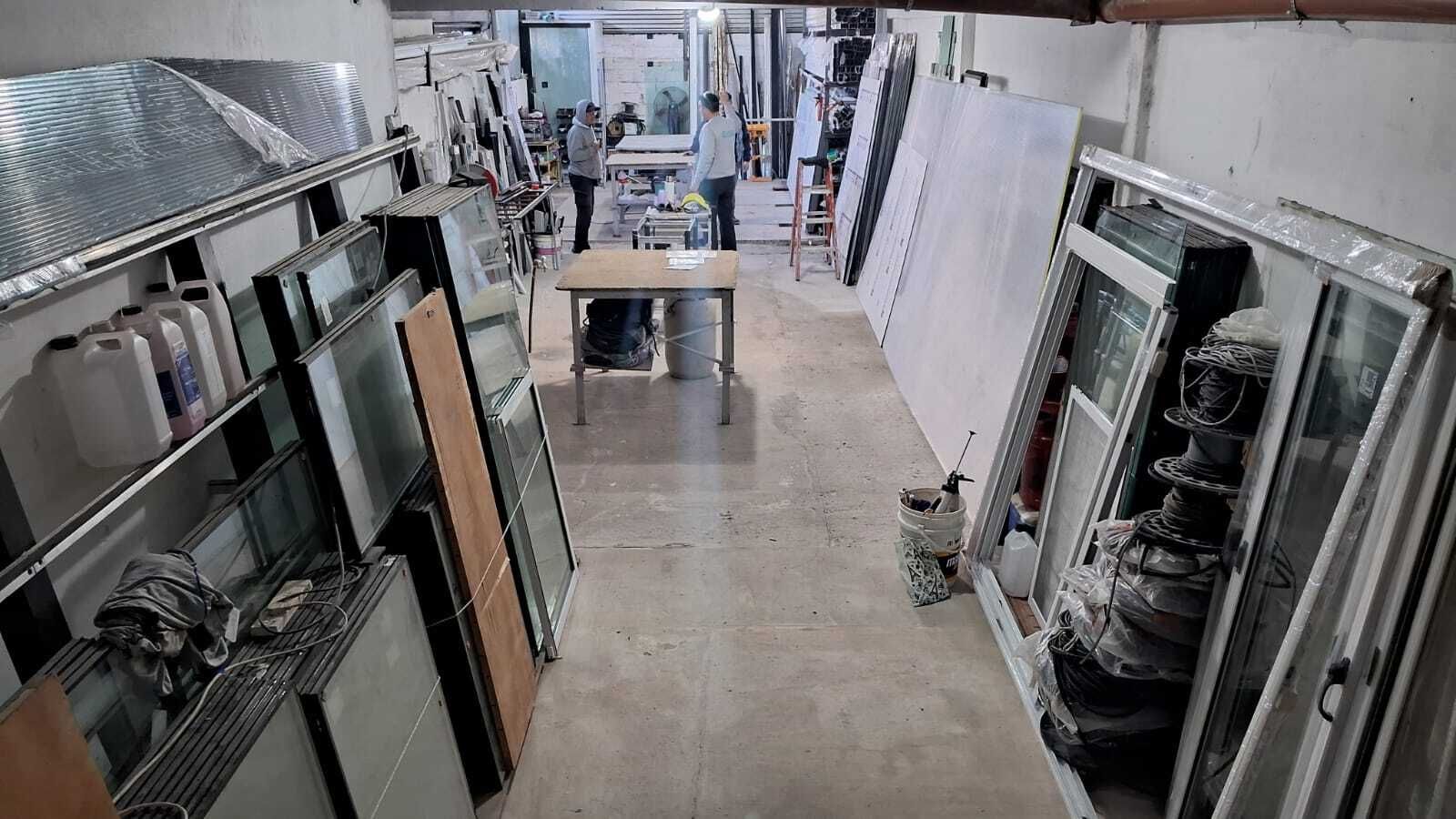 Un taller con ventanas, puertas y materiales. Dos personas están de pie junto a una mesa; estantes cubren las paredes.