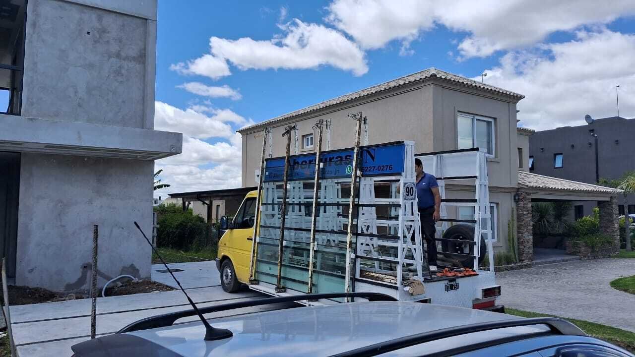 Furgoneta con grandes paneles de vidrio; trabajador en escalera, junto a una casa en construcción. Cielo azul, día soleado.