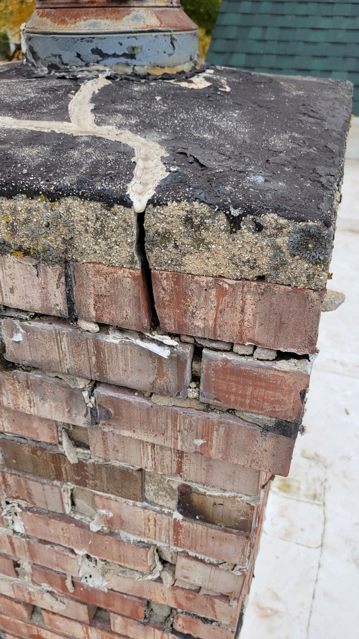 Cracked brick chimney with deteriorating cap. Brown bricks, black cap with white filler, weathered.