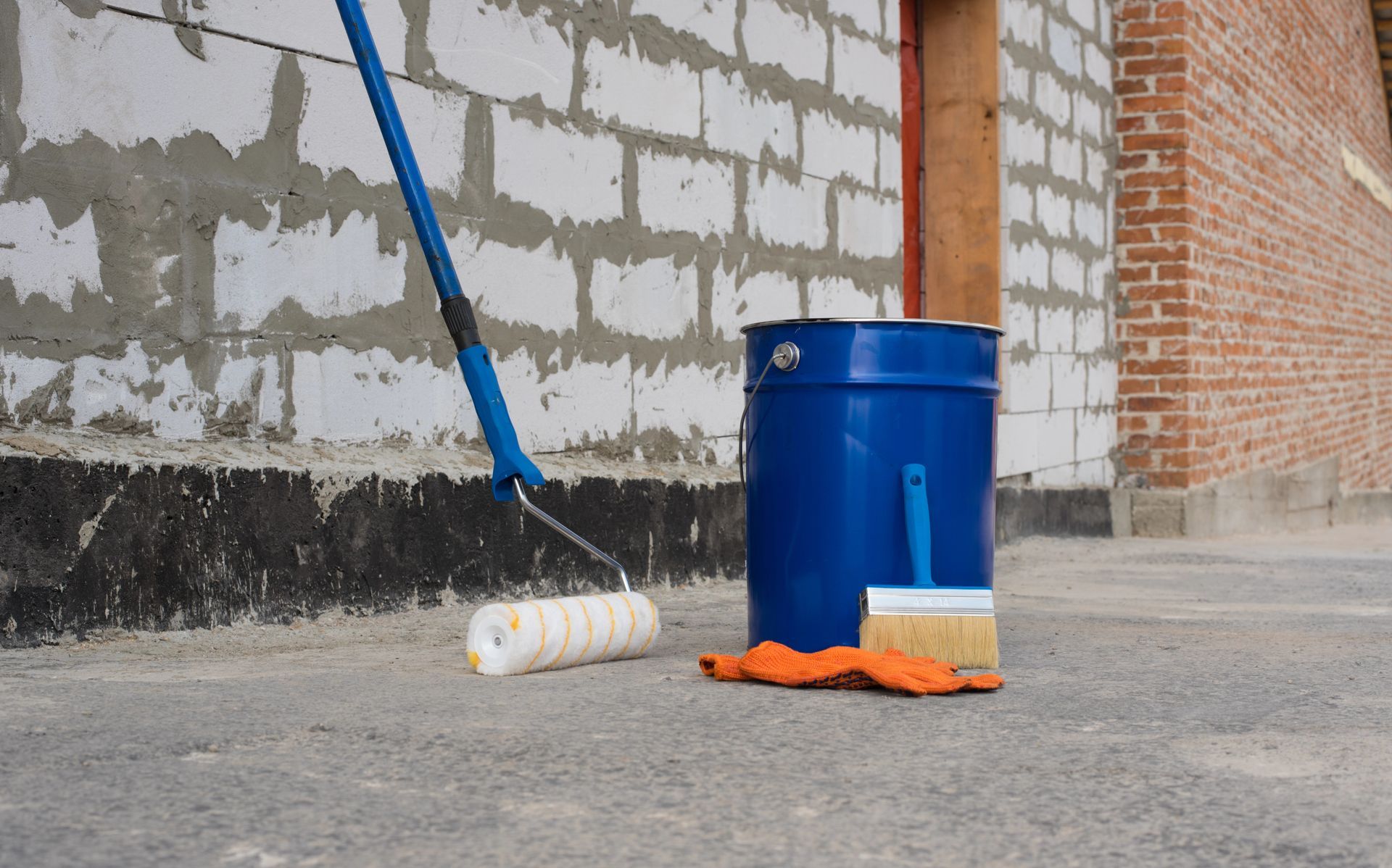 A bucket of paint , a roller and a brush are sitting on the ground in front of a brick wall.