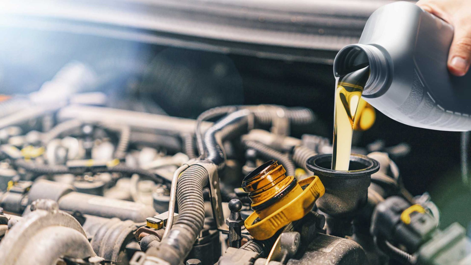 Motor oil being poured from a bottle into a car engine bay, with hoses and engine parts visible.