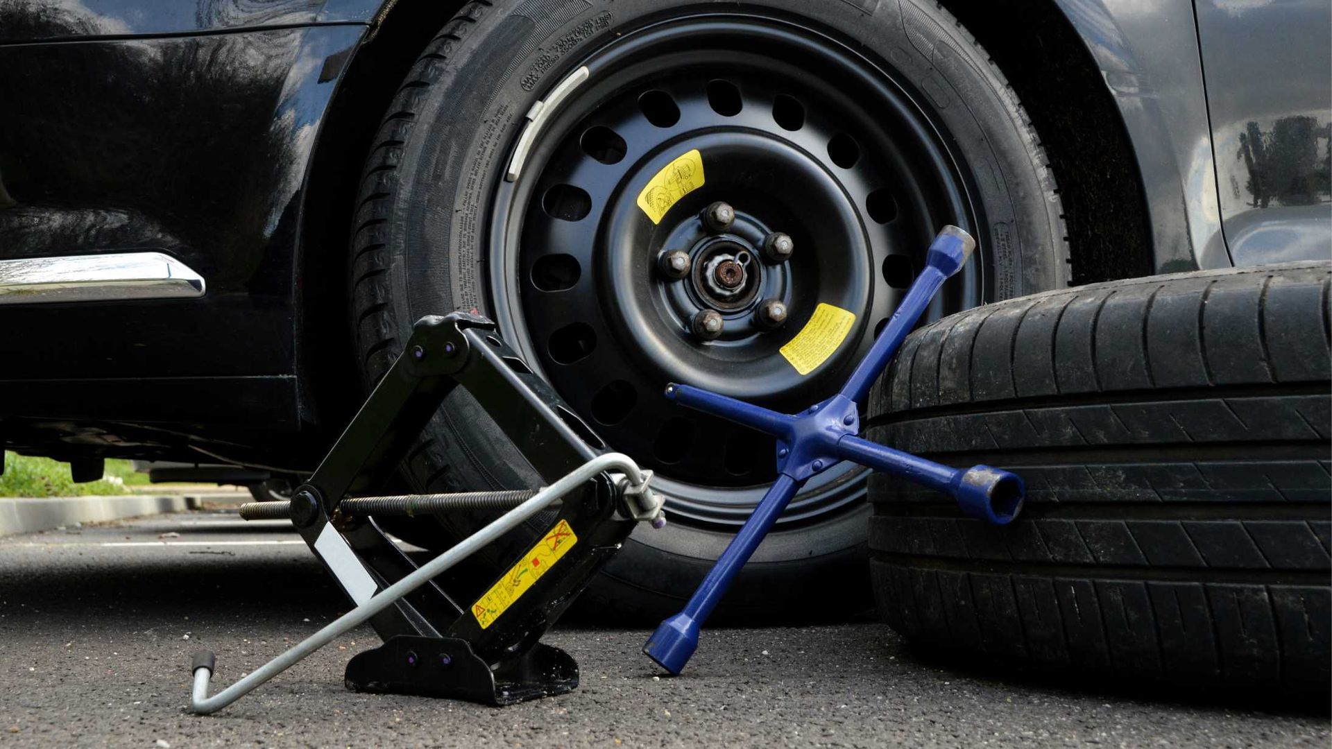 Car jack and lug wrench beside a car’s flat tire on the road, with a spare tire nearby