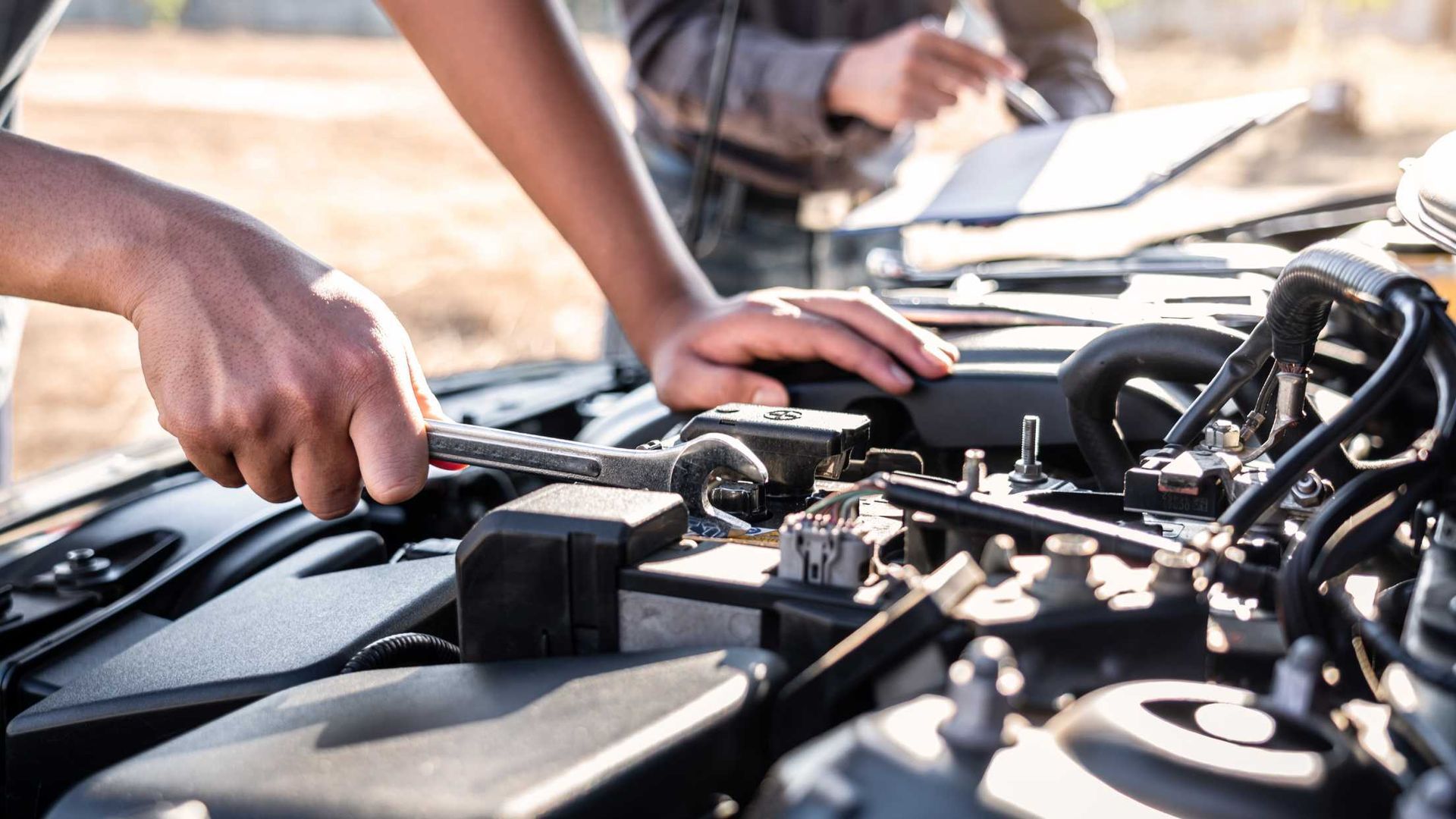 Two people working under a car hood, checking the engine in bright daylight.