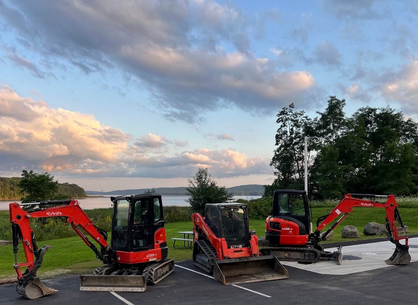 Three orange construction vehicles parked in front of a lake and grassy area under a cloudy sky.