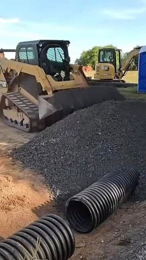 A yellow bulldozer and excavator at a construction site with a pile of gravel and a large corrugated pipe.
