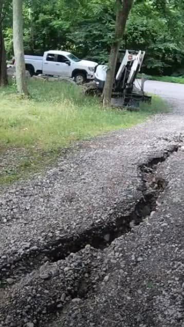 A cracked, gravel road with a white pickup truck and a backhoe in the background, near trees.