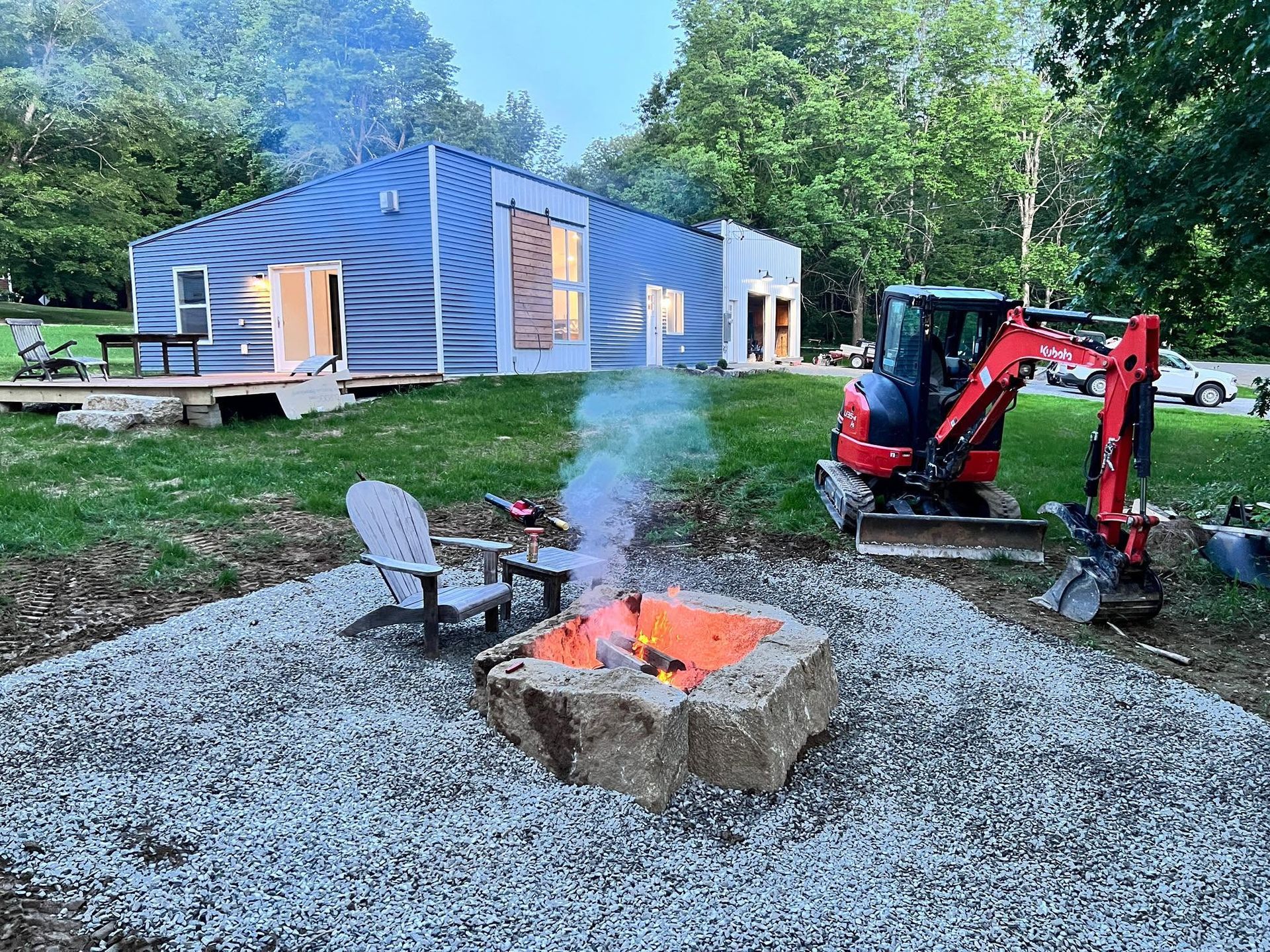 A modern blue house with a fire pit in front, a red excavator on the side. Two chairs sit by the fire, smoke rises.