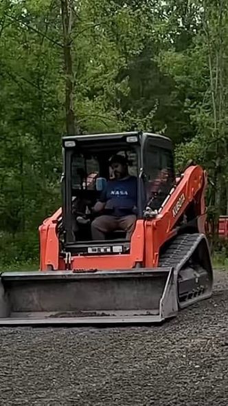 Man operating an orange Kubota track skid steer. He's in the cab, smiling. The machine is on gravel in a wooded area.