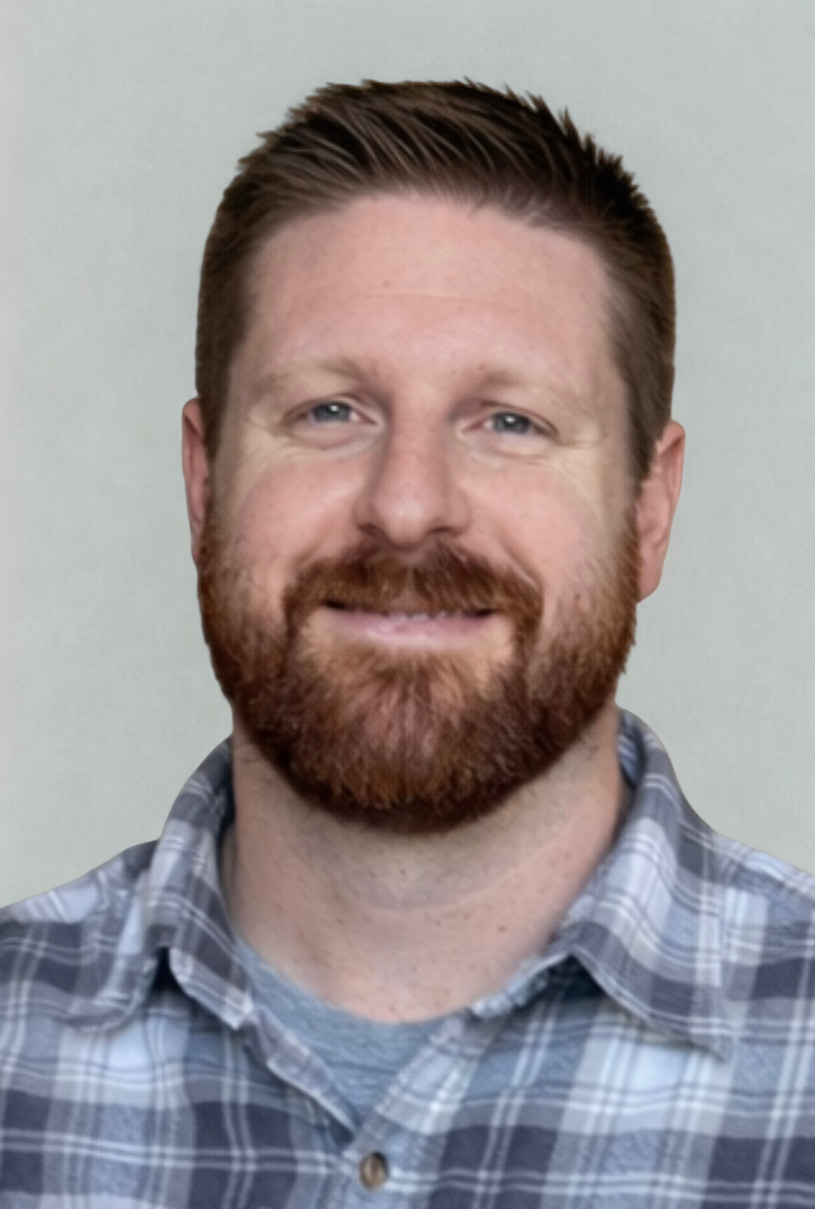 A person with a beard, wearing a gray and blue plaid shirt, smiles against a neutral background.