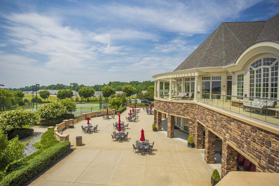 Outdoor patio with tables and umbrellas, next to a stone building with an open veranda. Tennis courts in the distance.