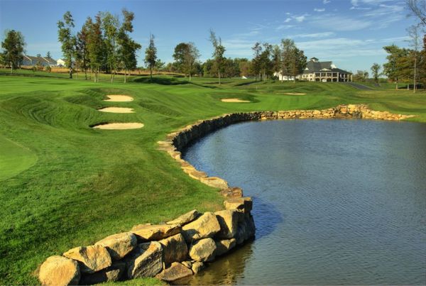 Golf course with green fairways, sand traps, water feature, and large stone wall; clear blue sky.