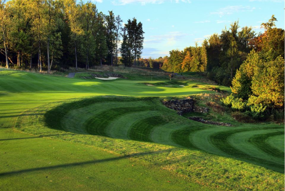 Golf course with striped green grass, trees, and a bunker under a blue sky.