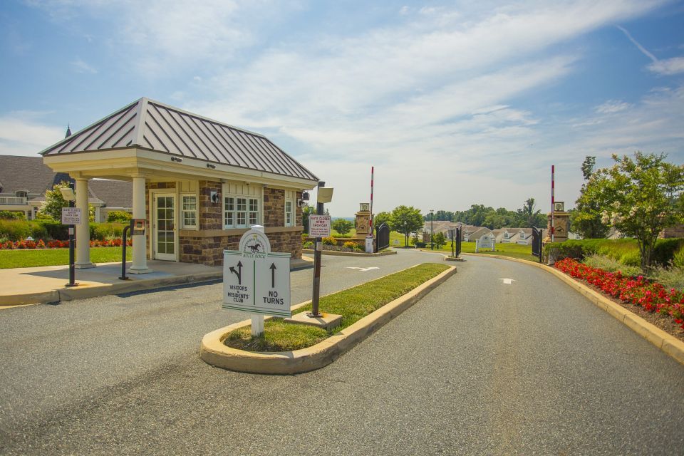 Entrance gate to a gated community with a guard house, asphalt road, and landscaping under a blue sky.