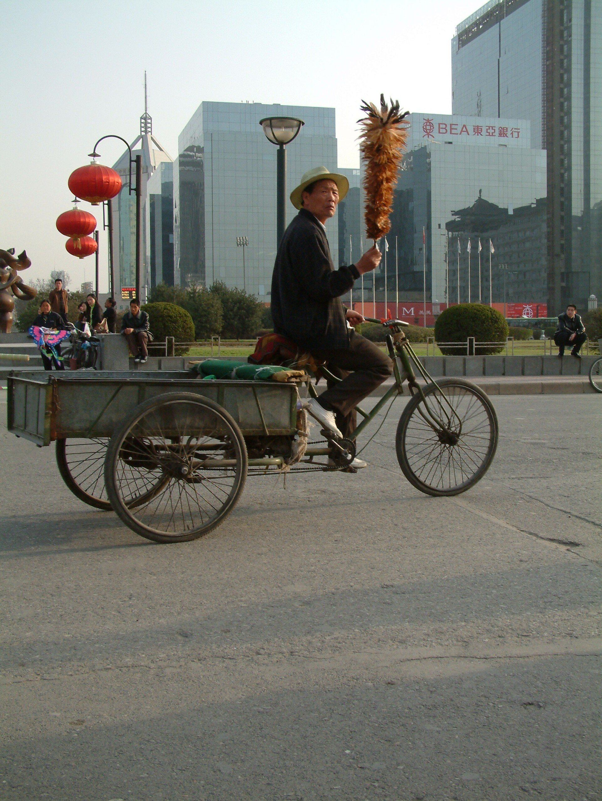 Een man op een driewieler met een karretje, met een plumeau in zijn hand, loopt over een stadsstraat voor moderne gebouwen.