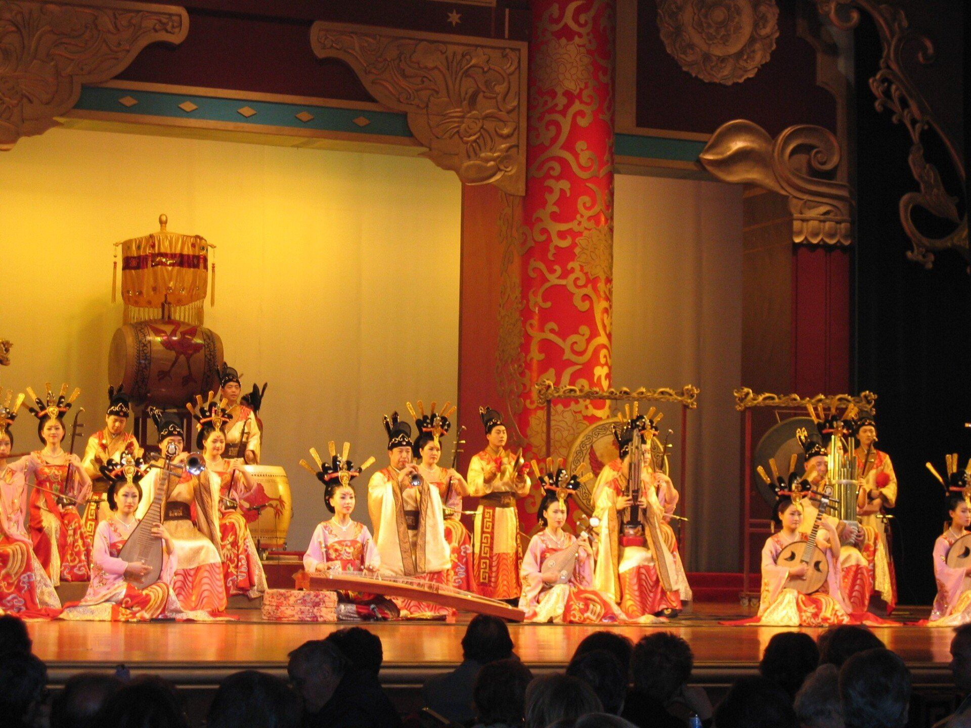 Toneelvoorstelling met muzikanten in traditionele Chinese kostuums, tegen een rijk versierd rood en goudkleurig decor.