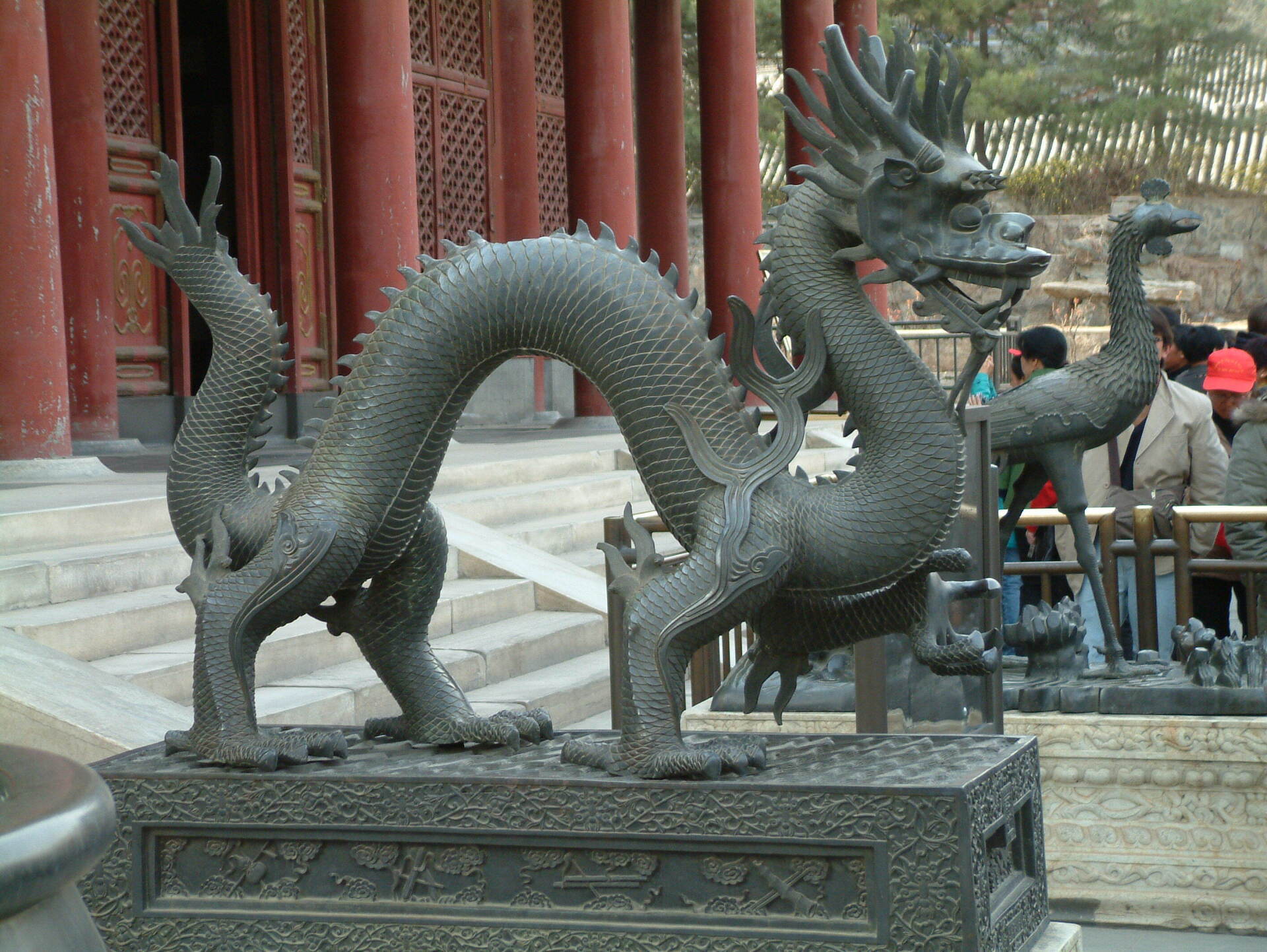 Bronzen drakenbeeld op een decoratieve sokkel, voor een gebouw met rode zuilen.