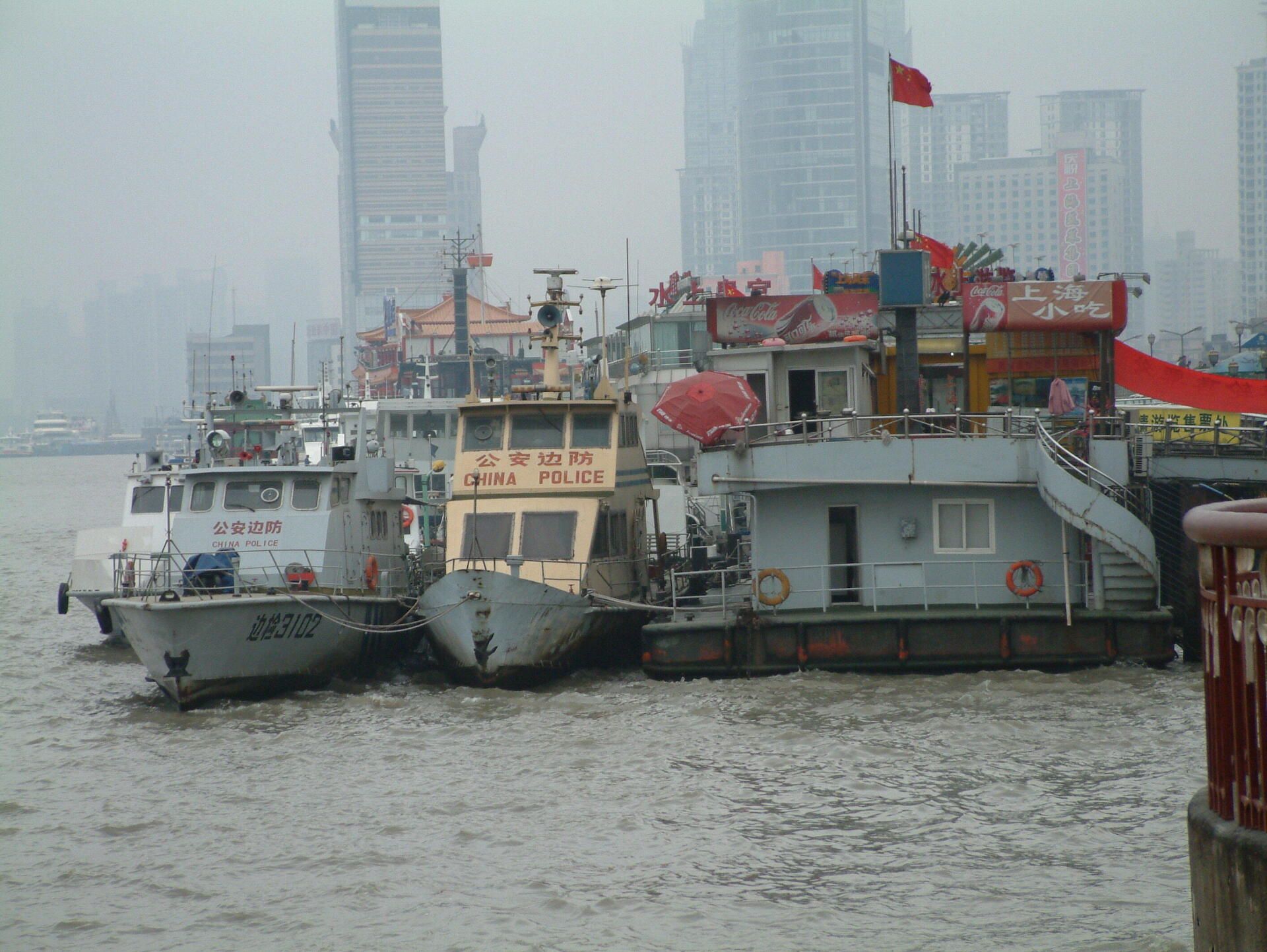 Boten aangemeerd in een haven, stadsgebouwen op de achtergrond, bewolkte dag.